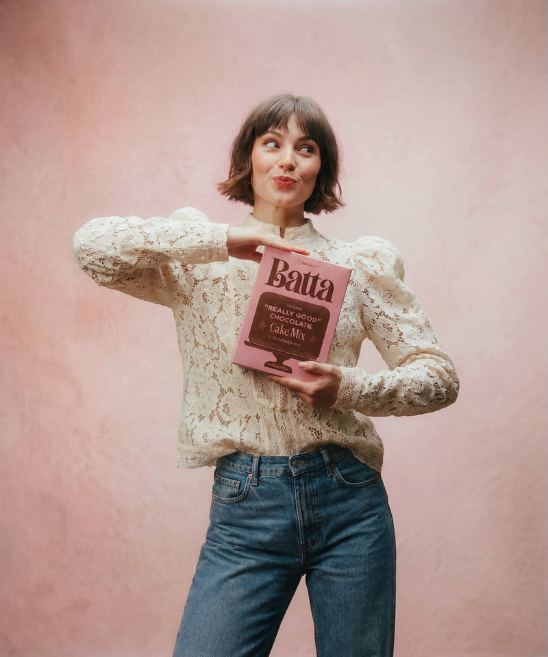 Woman holding a pink cake mix box against a pink background. Batta all-natural cake and frosting mixes - gourmet gifts for bakers, moms, foodies