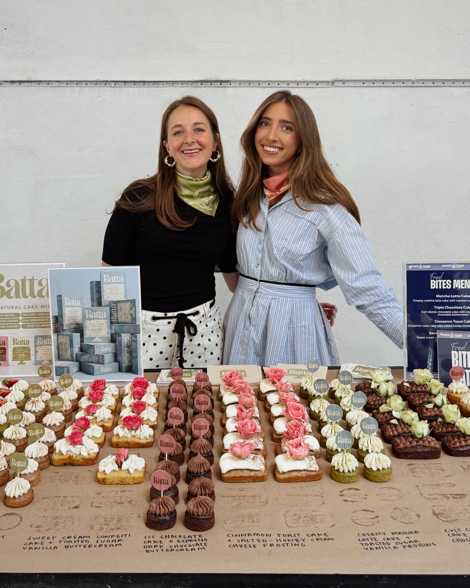 Two women standing next to a display of small cakes on a table. Batta all-natural cake and frosting mixes - gourmet gifts for bakers, moms, foodies